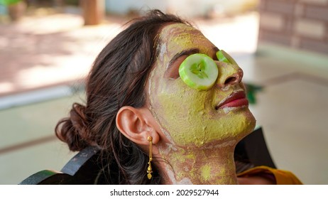 A Beautiful Woman With Facial Clay Mask On Face And Fresh Cucumber On Eyes Relaxing At Home. Portrait Of Beautiful Spa Girl. Beauty, Healthcare. Multani Mitti And Green Face Pack In India