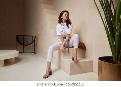Beautiful woman fashion model brunette hair tanned skin wear white overalls button suit sandals high heels accessory bag clothes style journey safari summer collection plant flowerpot wall stairs.