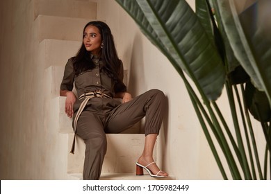 Beautiful woman fashion model brunette hair tanned skin wear green overalls button suit sandals high heels accessory clothes style journey safari summer collection plant flowerpot wall stairs.