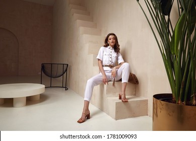 Beautiful woman fashion model brunette hair tanned skin wear white overalls button suit sandals high heels accessory bag clothes style journey safari summer collection plant flowerpot wall stairs.