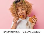 Beautiful woman feels very hungry eats chicken nuggets with ketchup wears dirty t shirt and protective helmet enjoys harmful fast food looks away isolated over pink background. Unhealthy eating