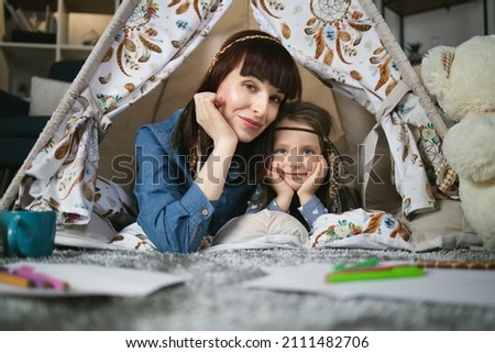 Beautiful woman and female kid in indian headbands smiling and looking at camera while lying together inside toy wigwam. Portrait of playful caucasian family.