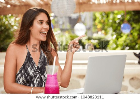 Beautiful woman freelancer with long hairstyle works on laptop computer, does remote job, indicates happily somewhere while sits in cozy cafeteria with fresh cocktail. People, leisure, recreation