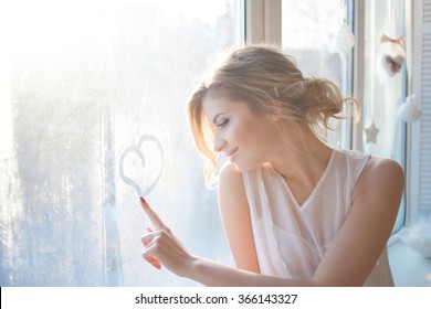 beautiful woman with fresh daily makeup and romantic wavy hairstyle, sitting at the windowsill, draws on glass