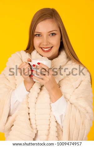 Beautiful woman in a fur winter jacket drinks tea over yellow background - cup of coffee