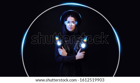 Beautiful woman in futuristic black dress over dark background. Gamer girl in glasses of virtual reality with controllers in hands. Augmented reality, game, hobby concept. VR. Blue neon light.