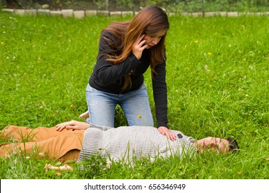 Beautiful woman giving first aid to a handsome young man and using her cellphone to call the ambulance, cardiopulmonary resuscitation, in a grass background