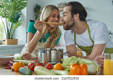 Beautiful woman giving her husband to taste a meal while both standing at the kitchen - Powered by Shutterstock