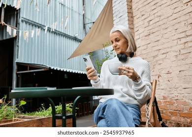 A beautiful woman with gray hair relaxes at a cafe, sipping coffee and using her smartphone. - Powered by Shutterstock