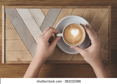 Beautiful Woman Hands Hold Ceramic White With Cappuccino Above Wooden Plate And Rustic Table. Milk Foam On Top In Heart Shape. Top View In Cafe Shop. Sale Presentation Concept.