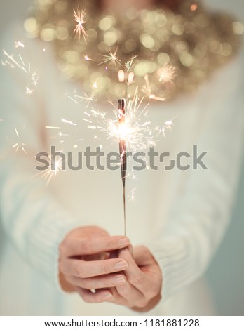 Beautiful woman hands holding sparkler lights, cozy, bright winter sweater with golden christmas garland, can be used as background