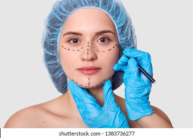 Beautiful Woman Has Marked Arrows Under Eyes. Lady Wants To Improve Her Appearance. Plastic Surgeon With Blue Gloves Draws Black Lines Before Face Lifting. Female With Medical Cap Against Grey Wall.