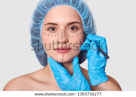 Beautiful woman has marked arrows under eyes. Lady wants to improve her appearance. Plastic surgeon with blue gloves draws black lines before face lifting. Female with medical cap against grey wall.