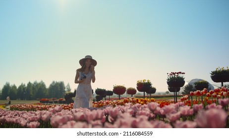 Beautiful woman in hat laughing to camera in spring flower garden. Girl with camera smiling in park with many flowers. Woman enjoying time in blooming flowers field. Pretty lady walking in meadow.  - Shutterstock ID 2116084559