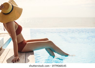 Beautiful woman in hat tanning on wooden pier at blue pool edge, enjoying summer vacation. Slim young female relaxing at tropical resort and sunbathing at swimming pool. Travel and Holidays