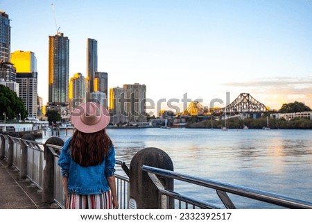 beautiful woman in a hat watching the sunset over brisbane city from kangaroo point, city reach boardwalk with amazing view of large skyscrapers by brisbane river, australia, queensland	