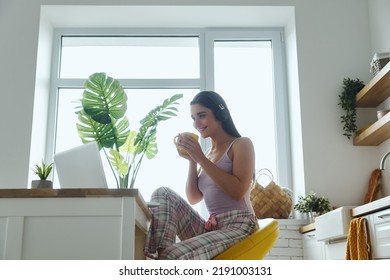 Beautiful Woman In Headphones Using Laptop And Drinking Coffee While Sitting At The Kitchen 