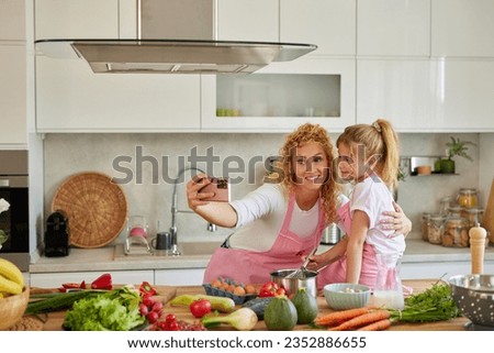 Beautiful woman and her cute daughter taking selfie in kitchen while preparing food