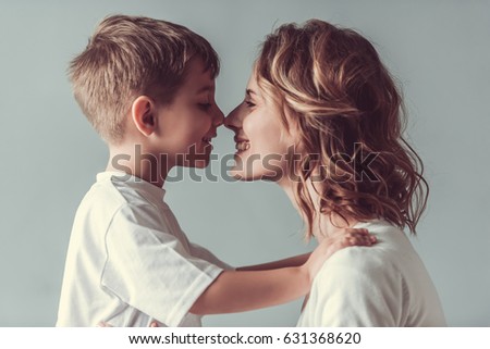 Beautiful woman and her cute little son are touching their noses and smiling, on gray background