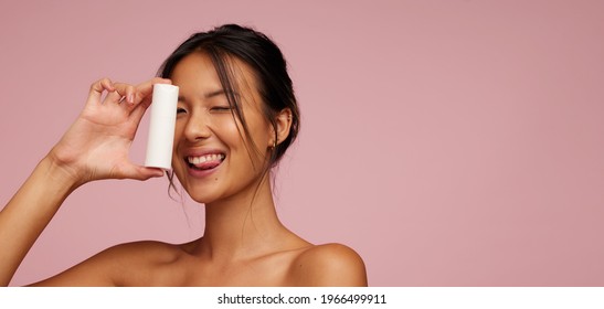 Beautiful Woman Holding A Beauty Product And Sticking Out Tongue Against Pink Background. Asian Female Model Showing A Skin Product.