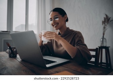 Beautiful woman holding coffee cup and looking at laptop while sitting at her working place - Powered by Shutterstock