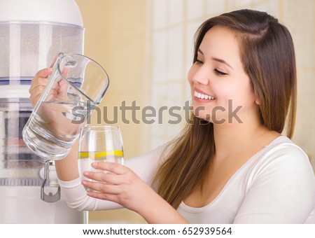 Beautiful woman holding a glass of water in one hand and a pitcher of water in her other hand, with a filter system of water purifier on a kitchen background
