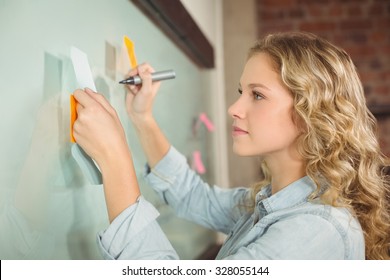 Beautiful Woman Holding Sticky Note While Writing On Glass Board In Creative Office