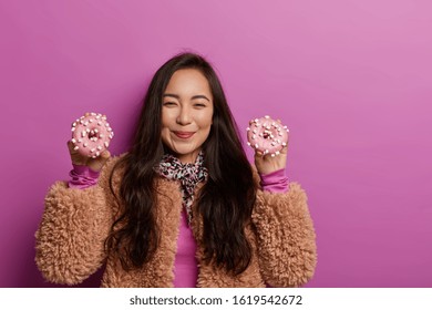 Beautiful Woman Holds Two Tasty Donuts In Both Hands, Has Glad Expression, Feels Temptation As Keeps To Diet, Wears Brown Coat, Isolated Over Lilac Wall, Blank Space. Junk Food, Unhealthy Desserts