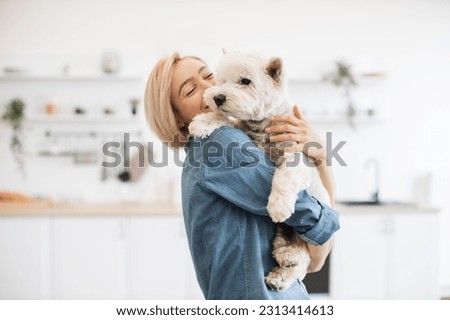 Beautiful woman in jeans shirt holding medium-sized dog on shoulder while standing in light room interior. Emotional female keeper finding joy in everyday interaction with furry friend indoors.