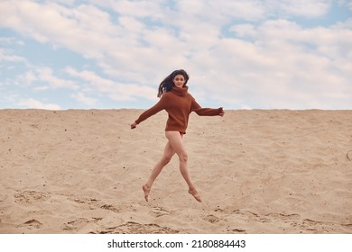 Beautiful Woman Jump Up With Windy Hair On Sandy Beach. Stylish Young Sexy Female In Knitted Sweater And Bare Legs Relaxing On Coast.