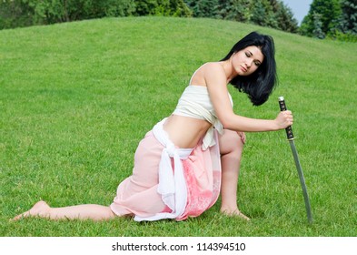 Beautiful Woman Kneeling On The Grass Posing Leaning Forwards On The Point Of A Sword