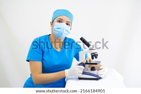 beautiful woman lab technician wearing uniform and blue surgical cap, surgical protection, analyzing samples with the microscope, on white background