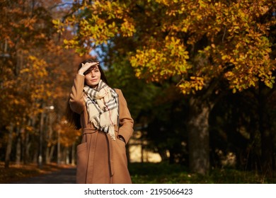 a beautiful woman in a light coat looks into the distance and covered her face with her hand from the sun on a walkway in an autumn park with fallen leaves. walk in autumn