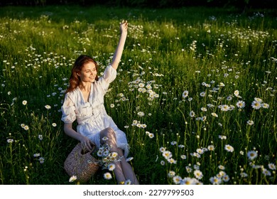 a beautiful woman in a light dress sits in a field of daisies with her hands raised above her head in the rays of the setting sun - Shutterstock ID 2279799503