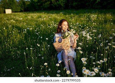 a beautiful woman in a light dress sits in a field of daisies with a wicker basket in her hands in the rays of the setting sun - Shutterstock ID 2279799535