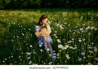 a beautiful woman in a light dress sits in a field of daisies with a wicker basket in her hands in the rays of the setting sun - Shutterstock ID 2279799551