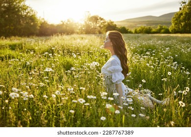 a beautiful woman in a light dress sits in a chamomile field against the backdrop of the setting sun and enjoys nature