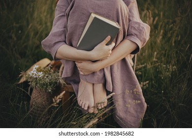 Beautiful woman in linen dress sitting barefoot on rustic chair with book in summer meadow, close up. Young female holding book and relaxing in countryside. Atmospheric calm moment
