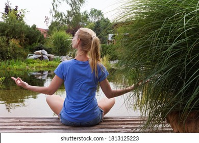 Beautiful Woman With Long Blond Hair Sitting In Zen Home Garden In Front Of Lake And Meditating. 