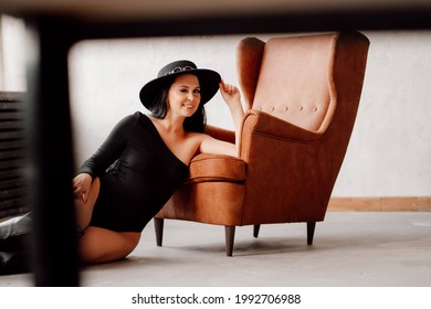 Beautiful Woman With Long Dark Hair In A Black Bodysuit And Hat On The Floor Of The Leather Chair. Country Style. Sexy Underwear And Clothes For The Theme Party.