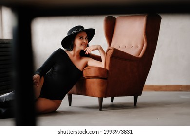 Beautiful Woman With Long Dark Hair In A Black Bodysuit And Hat On The Floor Of The Leather Chair. Country Style. Sexy Underwear And Clothes For The Theme Party.