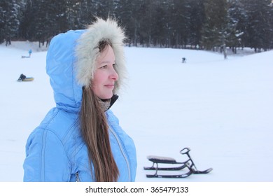 The beautiful woman with long hair in a blue jacket with a hood against snow and the wood in the winter