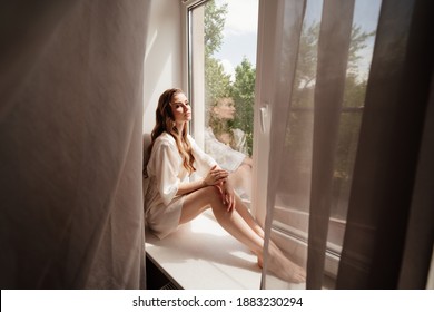 beautiful woman with long hair in white robe sits on windowsill. reflection in the glass. morning of bride before wedding. Natural beauty and professional make-up. beauticians and skin care products.