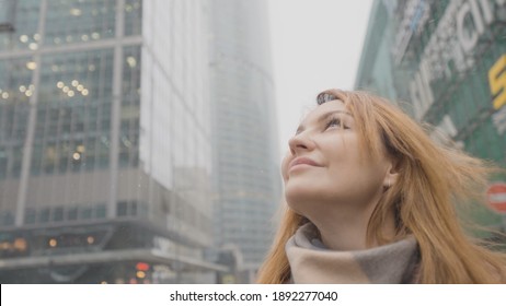 Beautiful Woman Looks Up At High-rise Business. Action. Woman With Confident And Determined Look Looks At Modern Skyscrapers. Motivated Woman Smiles Looking At Skyscrapers In City Center