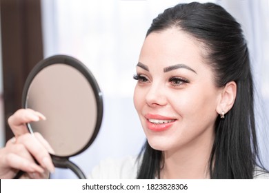 Beautiful Woman Looks In The Mirror And Smiles. The Girl Likes Her Appearance After Injecting Facial Rejuvenation At The Beautician. Lip Augmentation And Mesotherapy. Peeling And Cleansing Of The Face