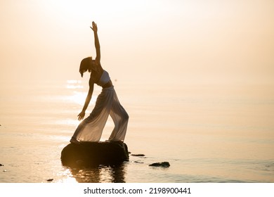 Beautiful Woman In Loose Clothing Practicing Yoga Or Pilates At Sunset Standing In Warrior Pose On Stone.