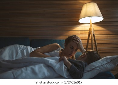 Beautiful Woman Lying With Little Boy In Bed And Telling Interesting Story Before Going To Sleep At Night. Night Shot Of Happy Smiling Mother Lying On Bed With Her Beautiful Baby Boy