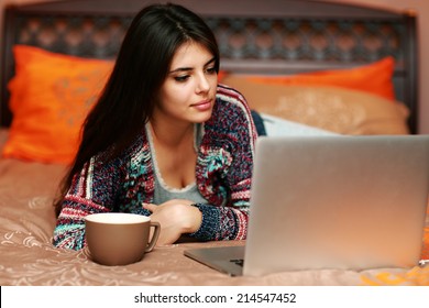 Beautiful woman lying on the bed with cup of coffee and looking at laptop - Powered by Shutterstock