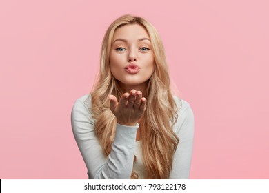 Beautiful woman with makeup and long blonde hair blows kiss, demonstrates her good feelings, says goodbye on distance, isolated over pink background. Young pretty female model makes air kiss.