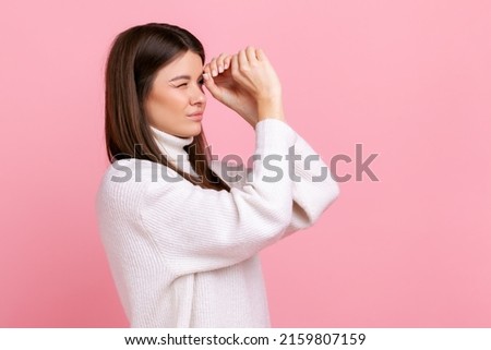 Beautiful woman making glasses shape, looking through binoculars gesture with attentive expression, wearing white casual style sweater. Indoor studio shot isolated on pink background.